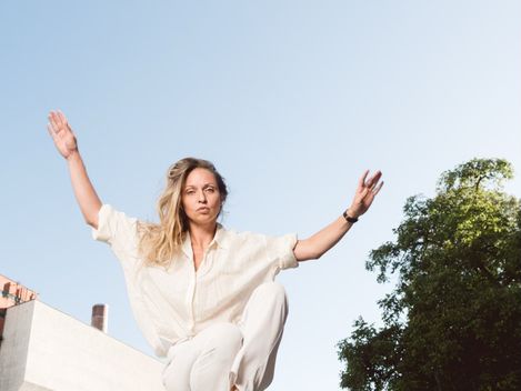 Person in heller Kleidung springt mit erhobenen Armen über ein Metallgeländer im Freien vor blauem Himmel