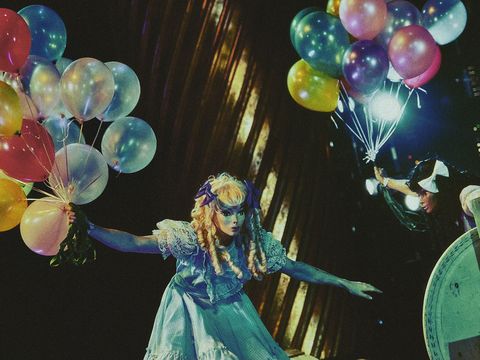 Two actresses wearing doll-like masks – one blonde, one brunette – both holding a bunch of balloons.