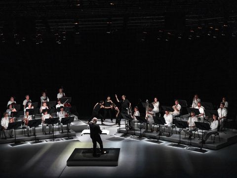Concert setting on a stage with a dark background. In the center, a small dance scene with four performers dressed in black. The musicians are positioned on the left and right, all wearing white shirts. In the foreground, the conductor can be seen from behind.