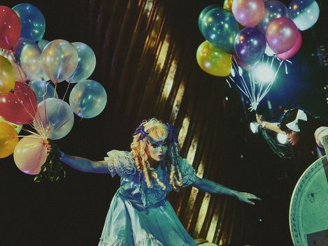 Two actresses wearing doll-like masks – one blonde, one brunette – both holding a bunch of balloons.