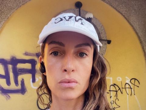 Person with long hair wearing a white cap standing in front of a yellow wall with black and white graffiti writing