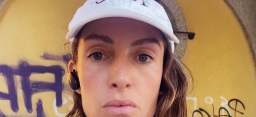 Person with long hair wearing a white cap standing in front of a yellow wall with black and white graffiti writing