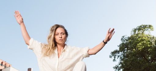 Person in heller Kleidung springt mit erhobenen Armen über ein Metallgeländer im Freien vor blauem Himmel