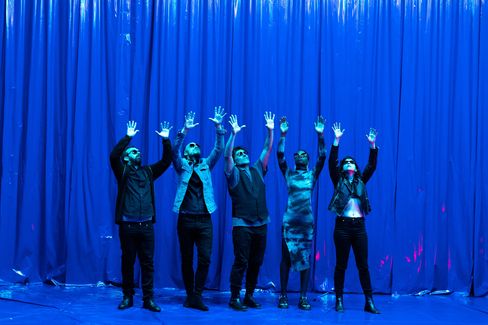 High emotions on a blue stage with a blue curtain and five performers raising their hands in the air.
