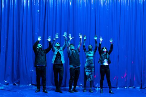 High emotions on a blue stage with a blue curtain and five performers raising their hands in the air.