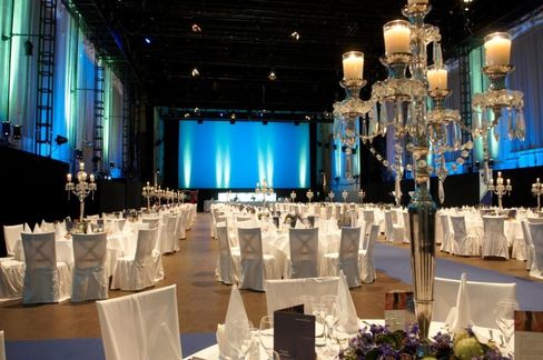 Atmospheric gala setting in Hall E of the MQ with beautifully laid tables and festive chairs with white covers, as well as richly decorated candlesticks resembling chandeliers. A screen can be seen in the background.