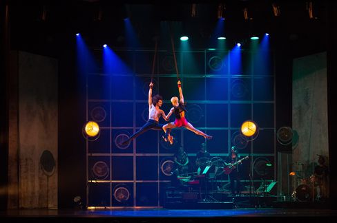 Stage scene from a musical show in which two performers are suspended in the air on a rope. A band consisting of three people can be seen in the background.