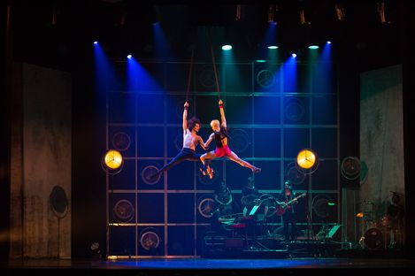 Stage scene from a musical show in which two performers are suspended in the air on a rope. A band consisting of three people can be seen in the background.
