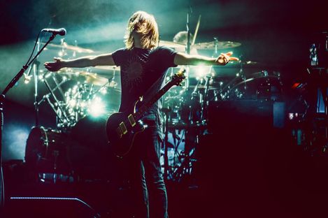 Concert stage with drums and microphones and a musician with long blond hair and an electric guitar, spreading his arms out to the right and left.
