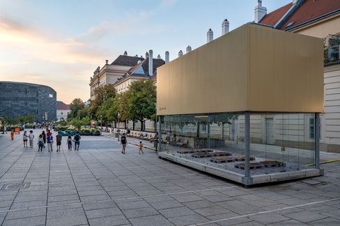 Large rectangular glass art installation with beige top on paved square in front of historic buildings and people in the background