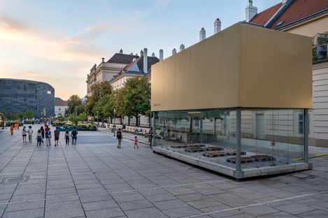 Large rectangular glass art installation with beige top on paved square in front of historic buildings and people in the background