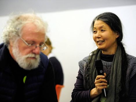 Two people indoors, one holding a microphone, the other sitting beside, with other people blurred in the background.