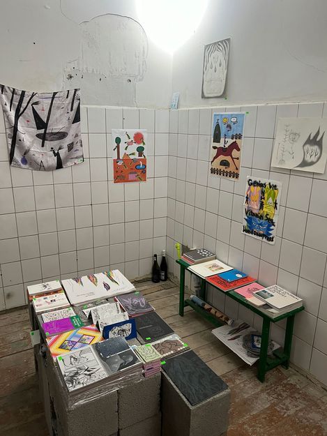 Kleiner Ausstellungsraum mit gefliesten Wänden, in dem Zeichnungen, Drucke und Bücher auf Tischen und an den Wänden präsentiert werden.