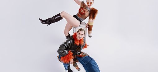 Zu sehen sind zwei Personen in ungewöhnlichem, kreativem Outfit, die Lust auf das Konzert machen.