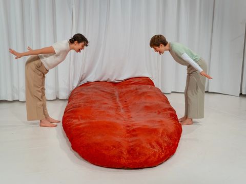 In the background: a white curtain. On stage: a huge red tongue. To the left and right of the tongue: two women sticking their tongues out at it.