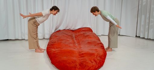 In the background: a white curtain. On stage: a huge red tongue. To the left and right of the tongue: two women sticking their tongues out at it.