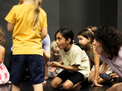 Kinder und die Workshopleitung kniet auf dem Tanzboden, den lächelnden Blick auf etwas Unsichtbares auf der linken Seite gerichtet.