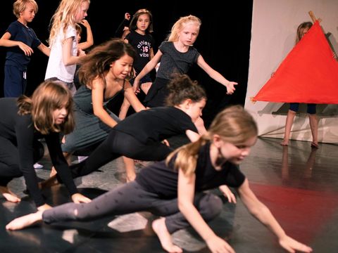 A group of children are crawling on the floor on their hands and knees. The children in the background are getting ready to do the same. On the right of the picture, a child is holding a large red fabric triangle.