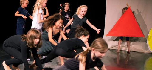 A group of children are crawling on the floor on their hands and knees. The children in the background are getting ready to do the same. On the right of the picture, a child is holding a large red fabric triangle.