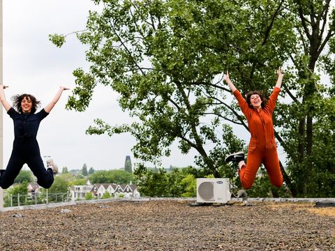 Two women in overalls on a rooftop. They are jumping into the air and are clearly enjoying it. In the background, one can see the roofs of a city.