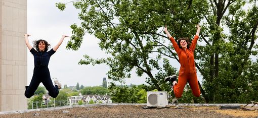 Two women in overalls on a rooftop. They are jumping into the air and are clearly enjoying it. In the background, one can see the roofs of a city.
