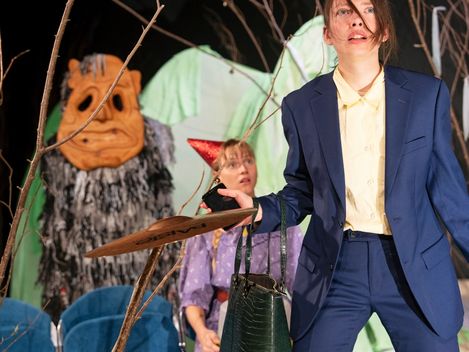 Zwei Frauen und ein freundliches Monster stehen im Wald. Die Frau im Vordergrund trägt einen blauen Anzug, die Frau im Hintergrund ein rotes Hütchen. Das Monster ist ein Zottelmonster.