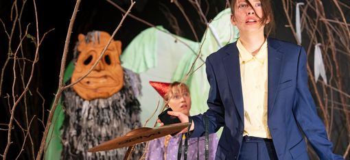 Zwei Frauen und ein freundliches Monster stehen im Wald. Die Frau im Vordergrund trägt einen blauen Anzug, die Frau im Hintergrund ein rotes Hütchen. Das Monster ist ein Zottelmonster.