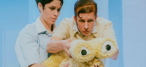 Two actresses, with artfully styled, immaculate hairstyles, are intently holding a cuddly creature with two large eyes and a red mouth.