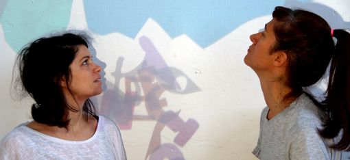 Two women in light-coloured T-shirts are looking at a wall covered in jumbled letters.