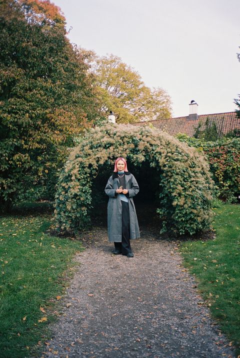 Porträt einer Person mit rosa Haaren, die auf einem Weg vor einem bewachsenen Torbogen in einem Garten steht.