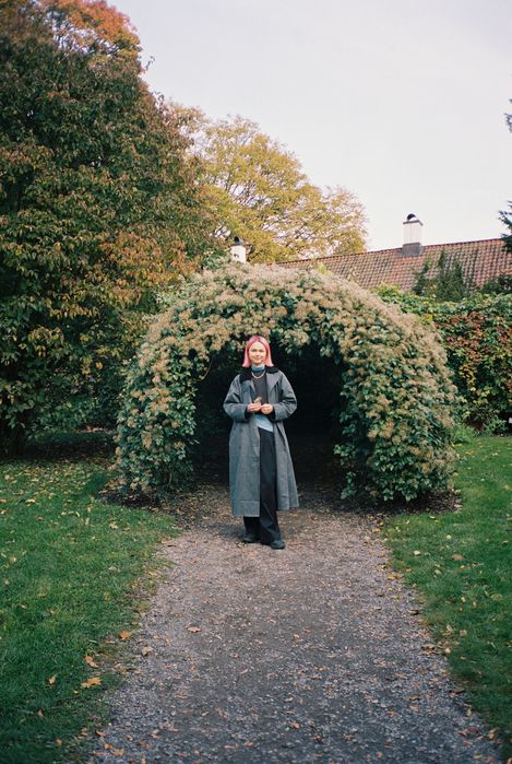Porträt einer Person mit rosa Haaren, die auf einem Weg vor einem bewachsenen Torbogen in einem Garten steht.